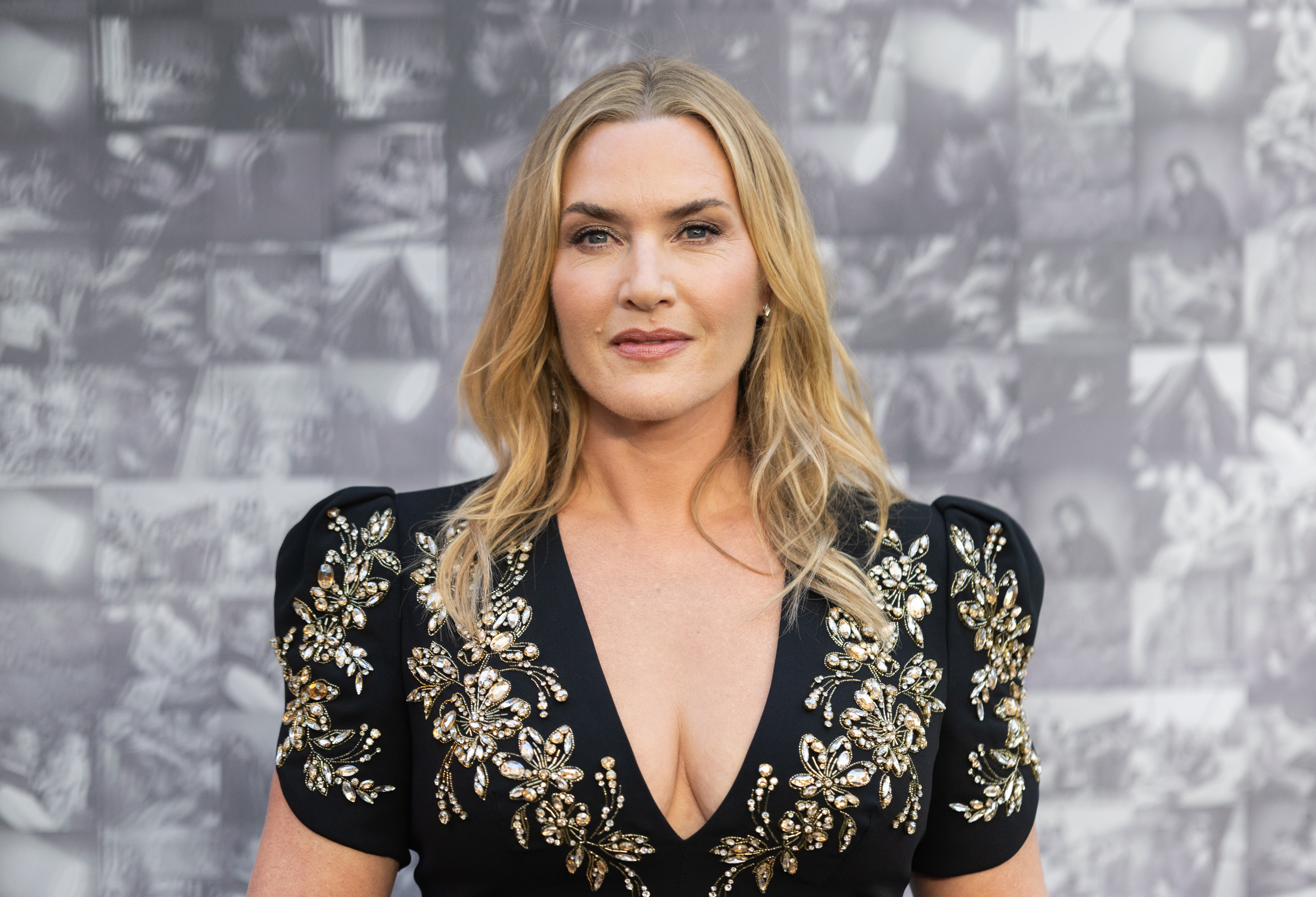 Kate Winslet at premiere for Lee. She wears black and gold dress and smiles at camera.