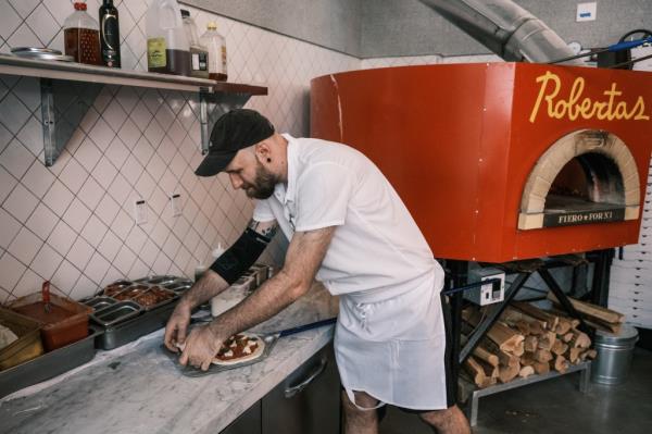 Jeff Walewski prepares a pizza atop a counter, with a red oven behind him