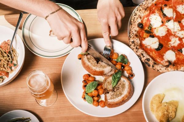 Hands holding a knife and fork to cut into slices of porchetta, arranged on a plate with small tomatoes and basil leaves.
