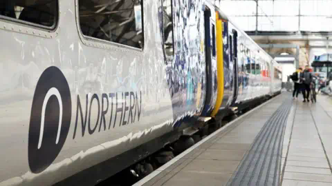 Northern A set of carriages on a Northern train running along a platform with the company sign seen on the outside of one of the cars. People can be seen at the end of platform waiting to alight.