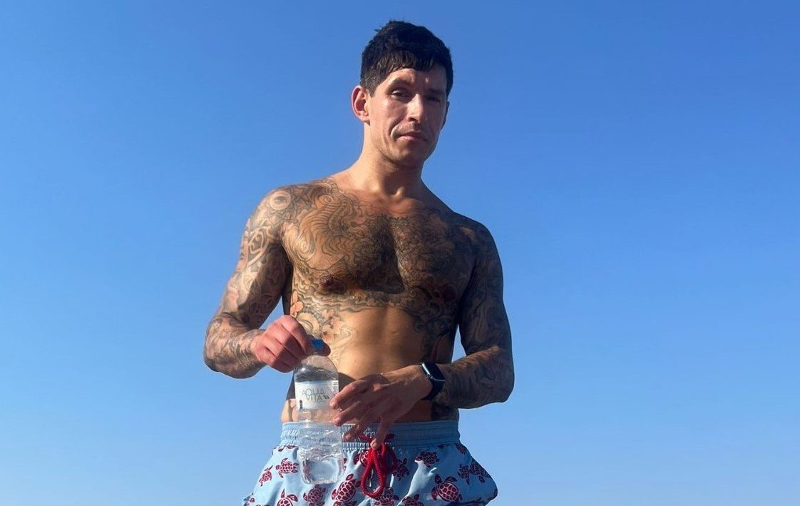 Marlon at the beach, with a blue sky behind him - he is topless and covered in tattoos