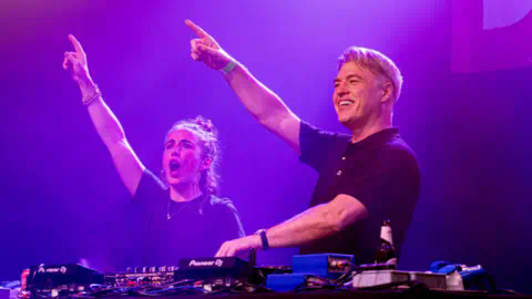 Day Fever Vicky McClure and Jonny Owen with one arm aloft while standing behind DJ decks