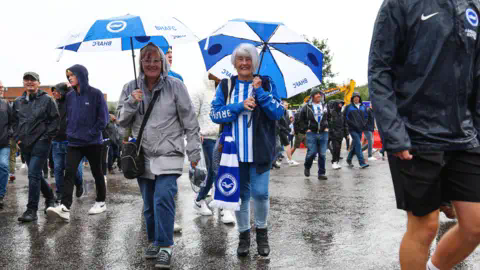 Getty Images Two women walking in the rain with umbrellas at the Amex stadium in Brighton