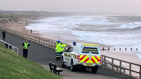 BBC Coastguard van at beach with grey skies and rough seas behind a line of police tape.