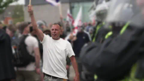 BBC Stephen Roughley穿着白色T恤，右手举着物品在骚乱中的照片。