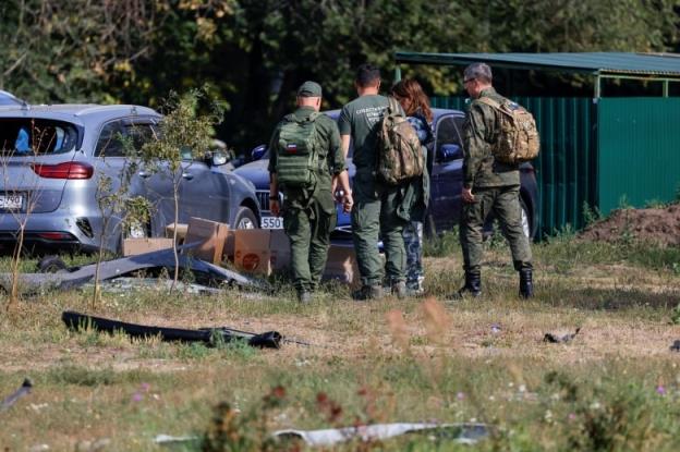 Several officials in uniform are shown in a grassy area near a damaged vehicle.