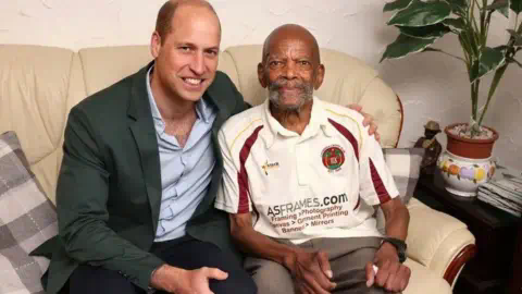 PA Media Prince William sits on a sofa with his arm round Alford Gardner, as they both smile into the camera. The Prince is wearing a green jacket and light blue buttoned shirt, while Mr Gardner is wearing a cricket top.