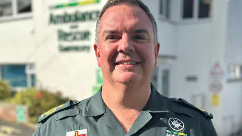 Guernsey Ambulance Service A man in a green ambulance service shirt smiles and looks towards us.