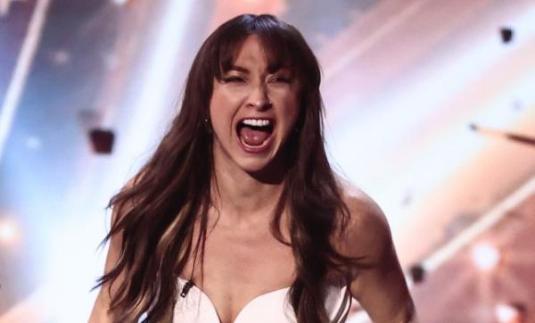 Sydnie Christmas celebrating after winning Britain's Got Talent. She is wearing a white jumpsuit. Her long brown hair is down and wavy. She appears to be shouting in celebration