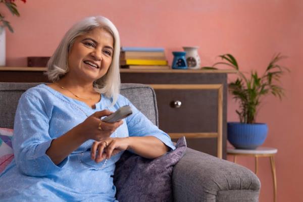 A middle-aged or older woman with gray hair sitting on the sofa in a colourful living room, smiling and pointing a television remote