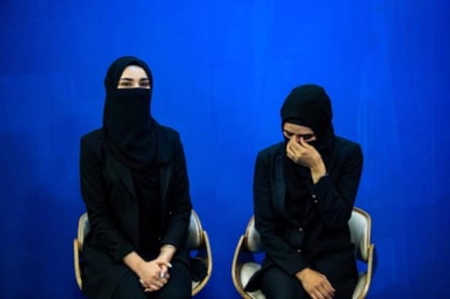 Two women sit in chairs wearing black hijabs and face coverings, against a blue background