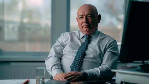Mick Sto<em></em>ney sitting behind a table in an office wearing a checked shirt and green tie