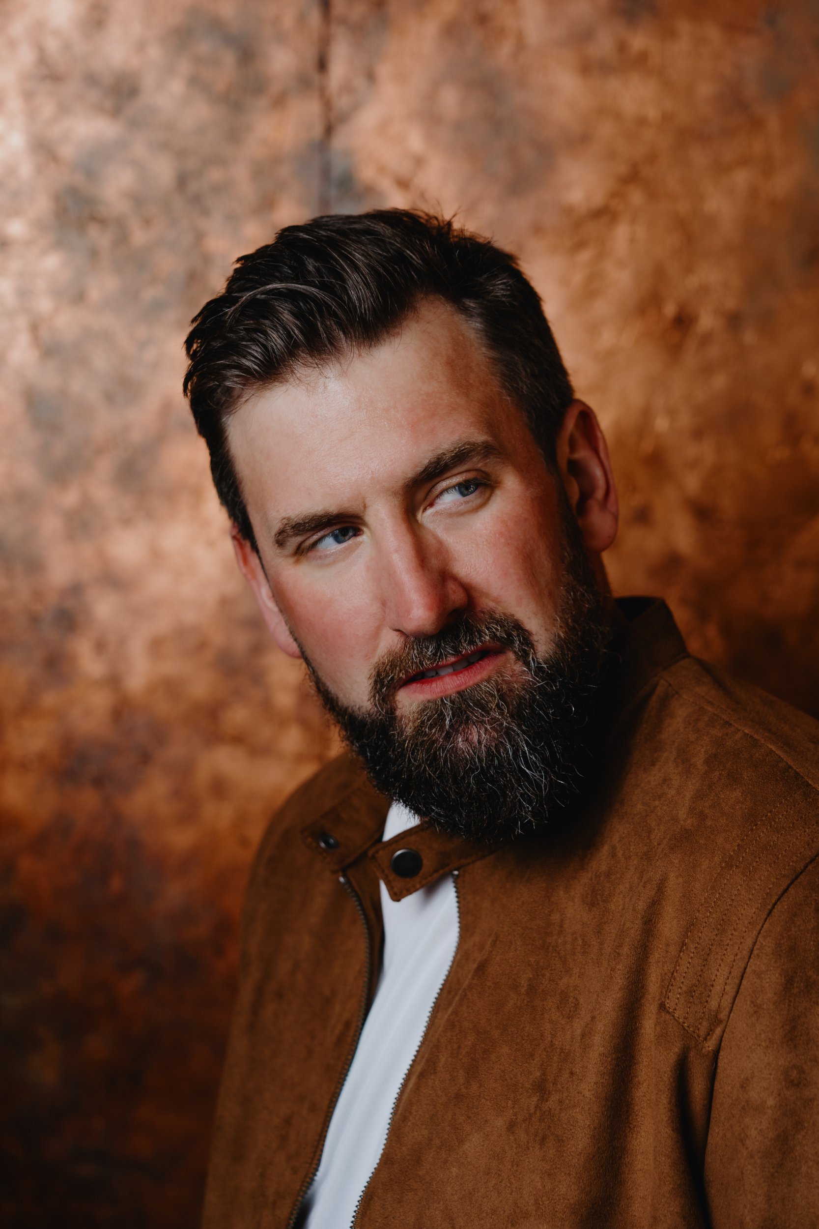 Garrett Millerick wearing a white t-shirt and tan jacket posing for promotional imagery