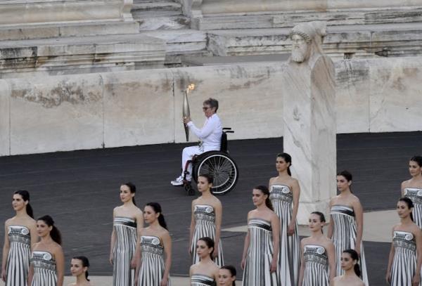 A white, older woman in a wheelchair carries a lit torch across the floor of a stadium