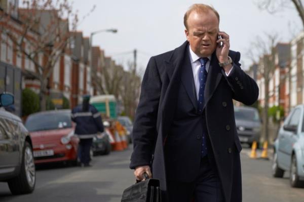 Toby Jones walking down a street speaking on the phone in a scene from Capital. 