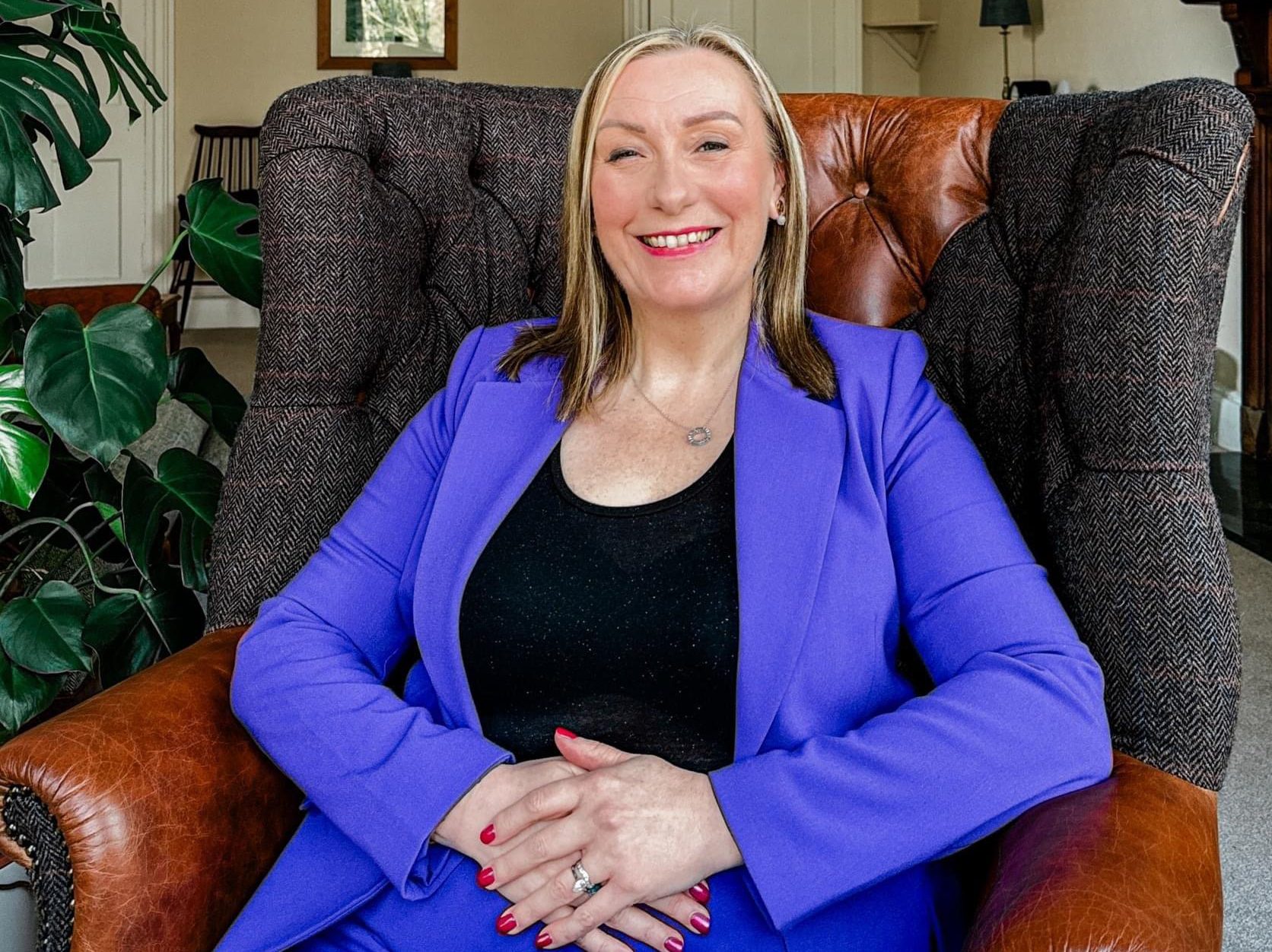 Julie sitting in an armchair with her hands folded, wearing a black top and purple blazer