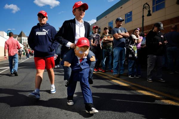 A young person wears a costume as he walks past hundreds of people waiting in line to attend a campaign rally with Republican presidential candidate, former President Donald Trump.