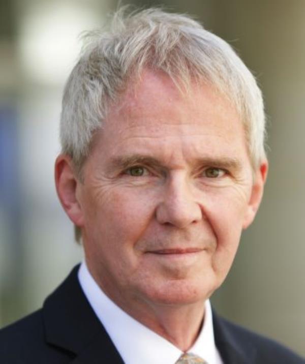 Portrait photo of a white man with short blond hair, wearing a black suit with a golden tie.