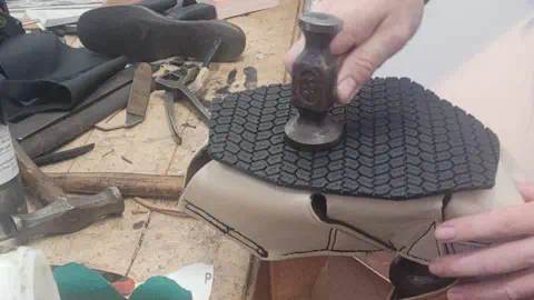 Stuart Moss Stuart Moss making the shoes for the camel. He is using a hammer to attach the black rubber soles to the boots, which are white and made of leather and memory foam.