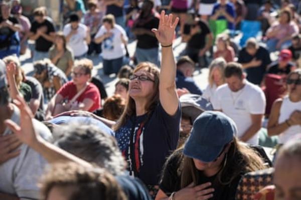 Crowd of people, with women in center raising hand in praise and wearing a US flag T-shirt.