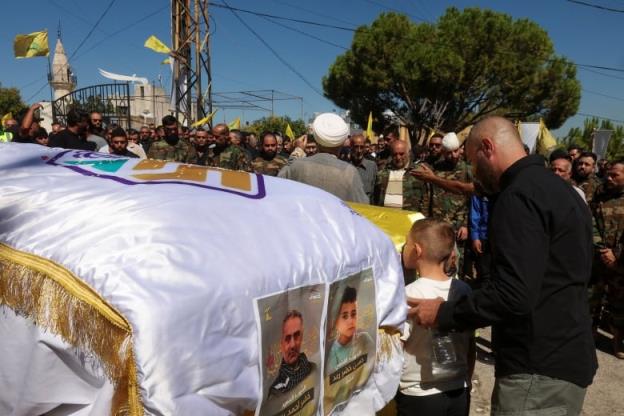 A small boy is shown kissing a coffin draped in an ornate covering. Dozens of people are standing nearby, including many in military uniforms.