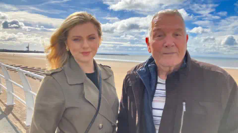 BBC A young woman with blonde hair standing alongside an older man with short grey hair on a promenade along a beachfront. There's golden sand behind them and the sea in the distance. The sky is blue with some clouds. In the left corner of the shot there is a large crane above a harbour. The woman is wearing a grey, buttoned front coat with a leather handbag strap across her front. The man is wearing a dark jacket with a blue top underneath and a white t-shirt. 