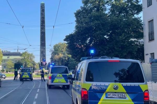 Police vehicles parked in Munich near the Nazi documentation Center and the Israeli Consulate General, with an obelisk in the background and tram tracks on the road.