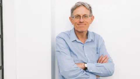 一位戴眼镜、穿着牛仔裤和蓝色敞开领衬衫的灰发男子,笑容满面,靠在白色墙壁上,右侧是一扇老式工业风格的窗户。