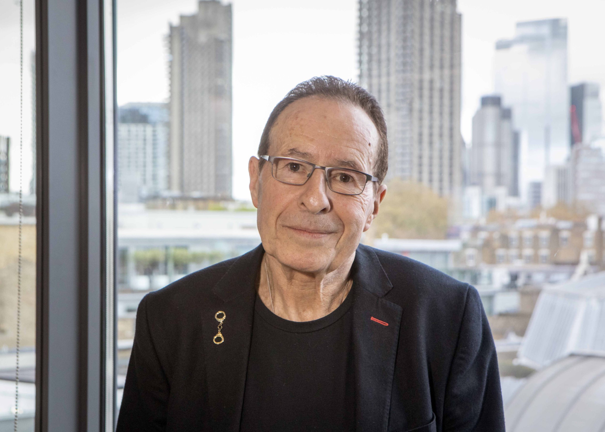 彼得坐在室内,背景是城市景观 - 他穿着黑色西装外套和T恤,戴着黑框眼镜
