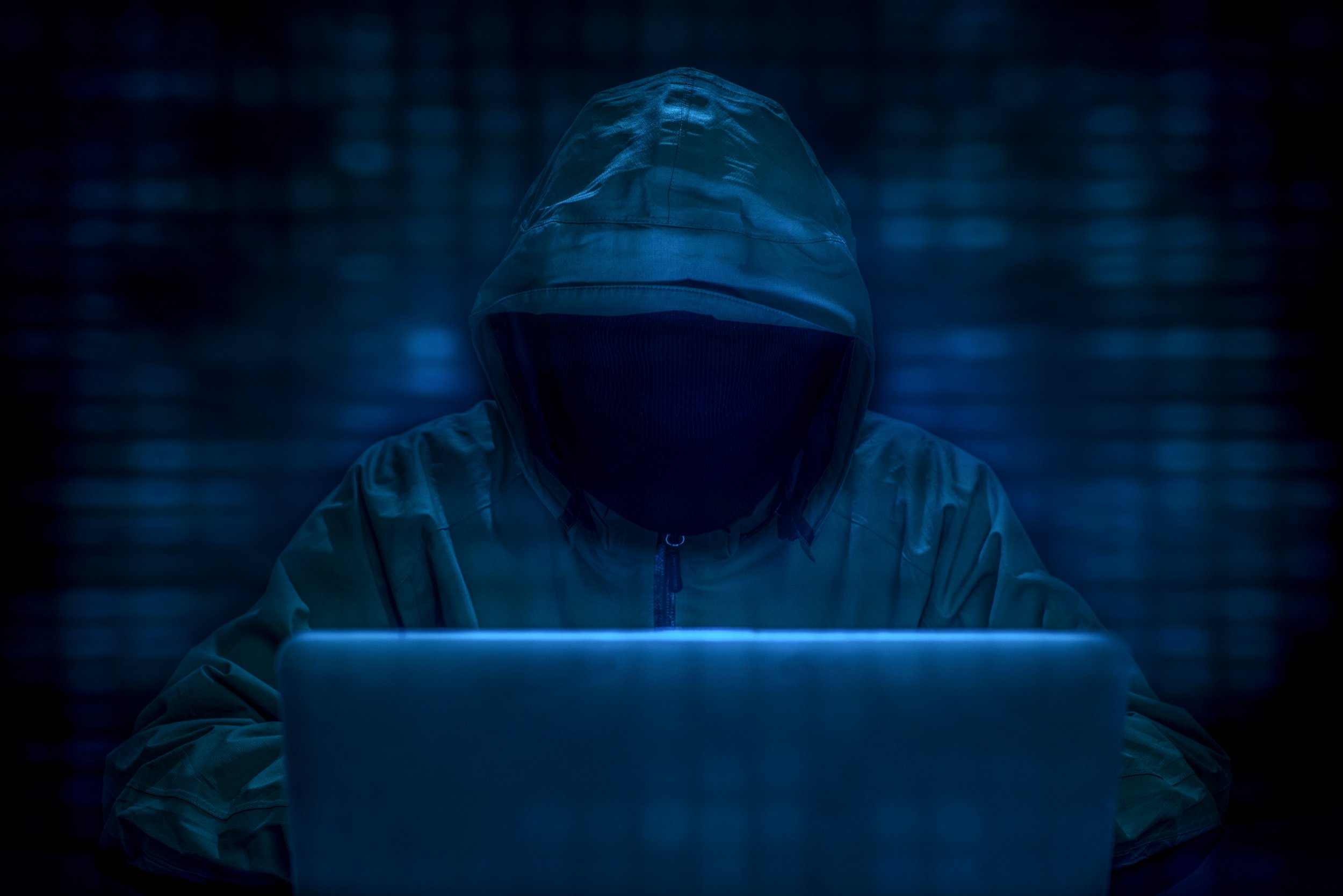 计算机黑客概念图,显示一个在黑暗房间里使用笔记本电脑的黑衣人