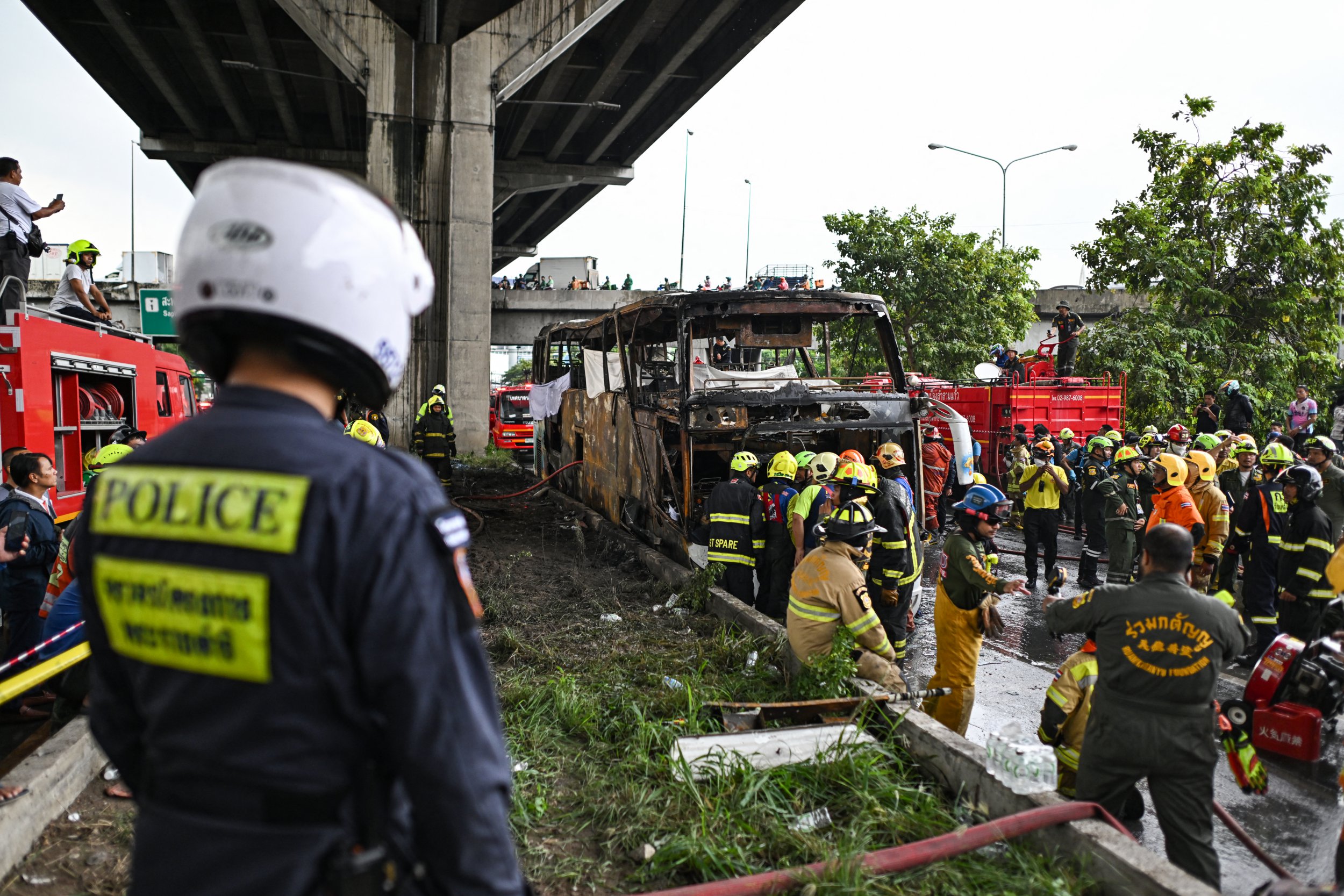 TOPSHOT - Firefighters and rescue workers stand next to a burnt-out bus that was carrying students and teachers on the outskirts of Bangkok, on October 1, 2024. A devastating fire tore through a Thai bus carrying 44 students and teachers on a school trip on October 1, 2024, officials said, with up to 25 feared dead. (Photo by Manan VATSYAYANA / AFP) (Photo by MANAN VATSYAYANA/AFP via Getty Images)