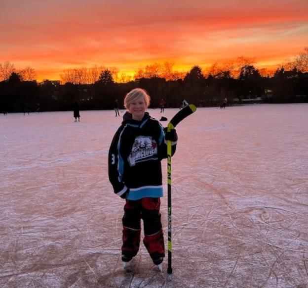 A smiling boy wearing a hockey jersey and holding a hockey stick stands in his skates on a frozen outdoor rink as the sun sets.