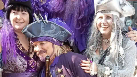 James Lacey Two women and a man dressed in pirate costumes. The man is wearing a purple tricorn hat and holding a replica pistol