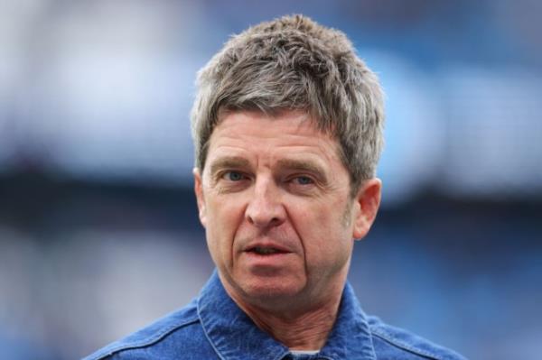 Noel Gallagher wearing a blue shirt standing in front of a blurred background