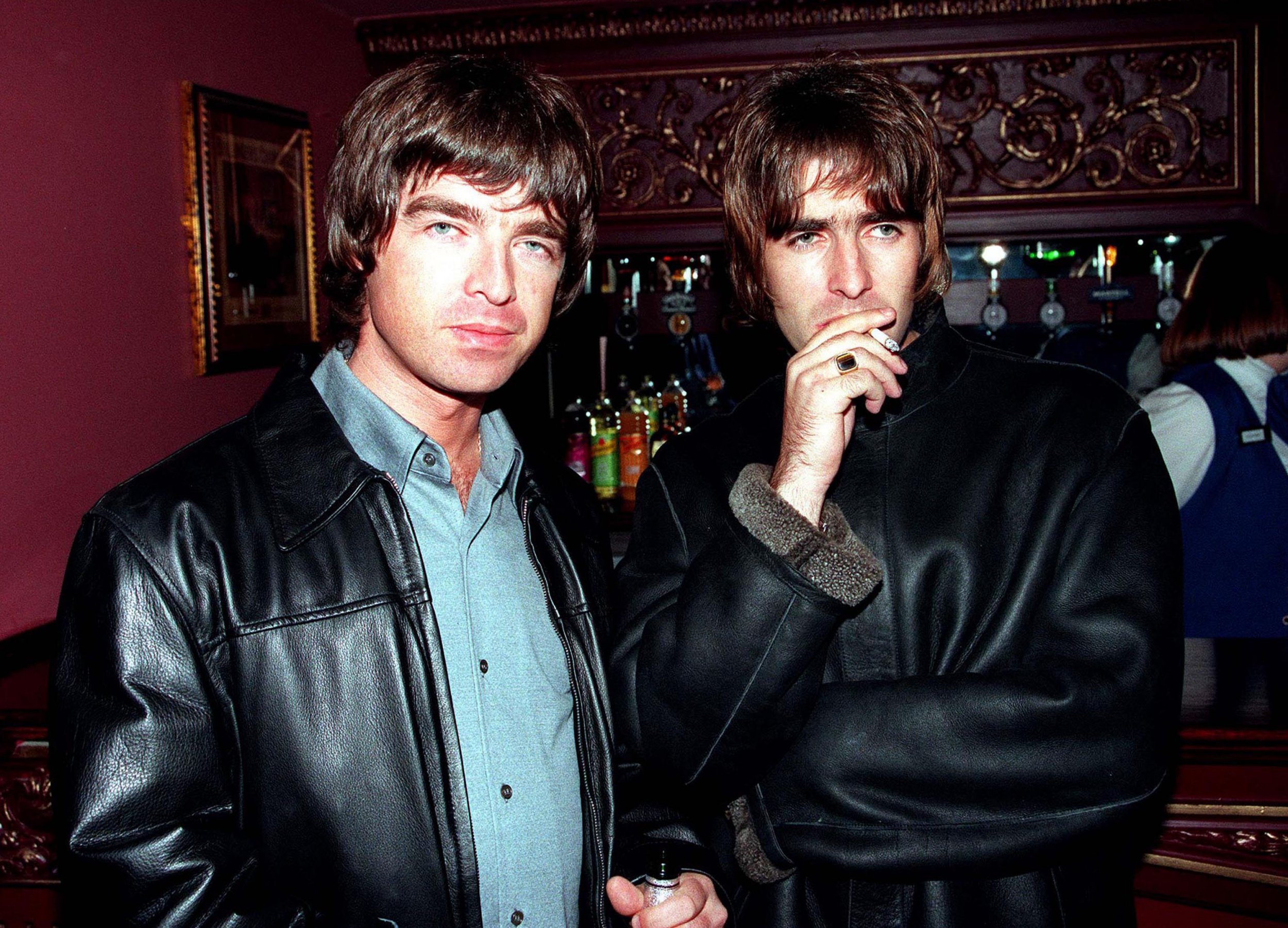 Liam Gallagher and brother Noel Gallagher standing next to each other in front of a bar. Liam is smoking a cigarette while Noel smiles at the camera