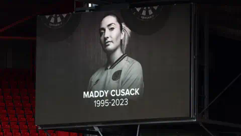 Getty Images 屏幕显示了前谢菲尔德联球员Maddy Cusack的纪念,当时谢菲尔德联和水晶宫之间的比赛正在进行。照片是一张黑白照片,Maddy穿着球衣,头发扎成马尾辫。下面写着:Maddy Cusack 1995-2023。