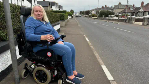 A woman in a motorised wheelchair looks off to the side of the camera. She has light blonde hair and is wearing a blue fleece top and blue jeans. She is at a bus stop on the side of a residential street. In the background you can see grey paving, homes and light traffic in the distance.