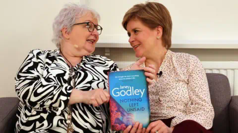PA media Janey Godley and Nicola Sturgeon, holding a copy of Ms Godley's book.